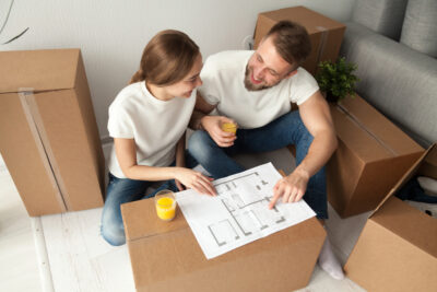 Happy couple discussing house plan looking at blueprint planning new home interior design, happy homeowners share furnishing remodeling ideas sitting on floor with moving boxes drinking orange juice