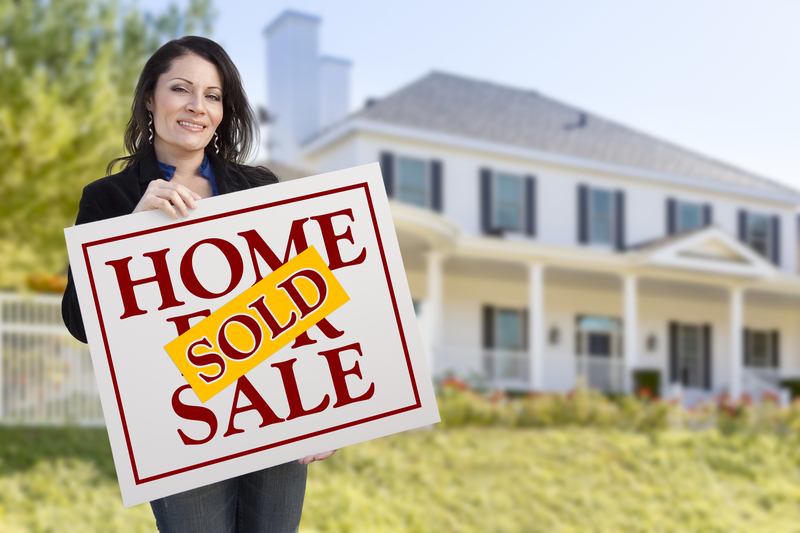 Smiling Woman Holding Sold Home For Sale Sign In Front of Beautiful House.